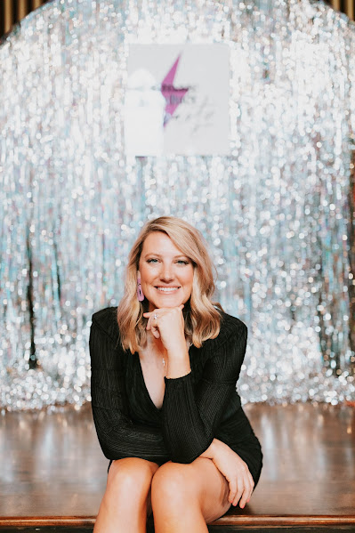 blonde woman sitting in black dress smiling silver sparkly background